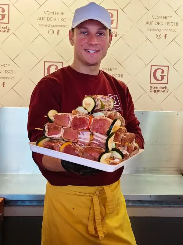 Herr Gutmann steht mit einem Teller angerichteter Grillspieße, lächelnd vor einer beigen Wand mit der Aufschrift: ,,Vom hof auf den Tisch!"